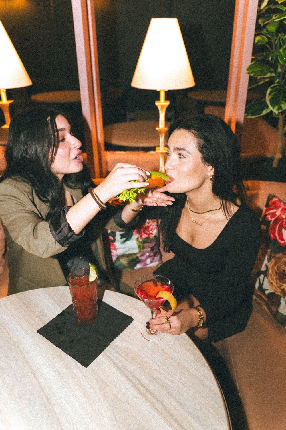 Two friends enjoying drinks and food at a dimly lit restaurant with table lamps and plants in the background.
