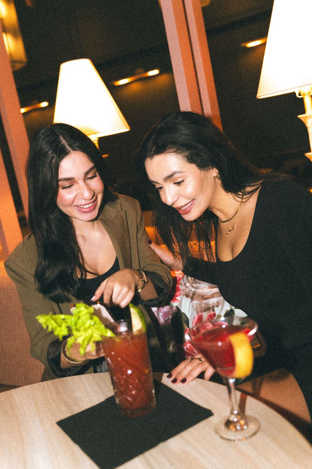 Two friends sharing laughter and colorful cocktails at a dimly lit restaurant with warm lighting and cozy atmosphere.