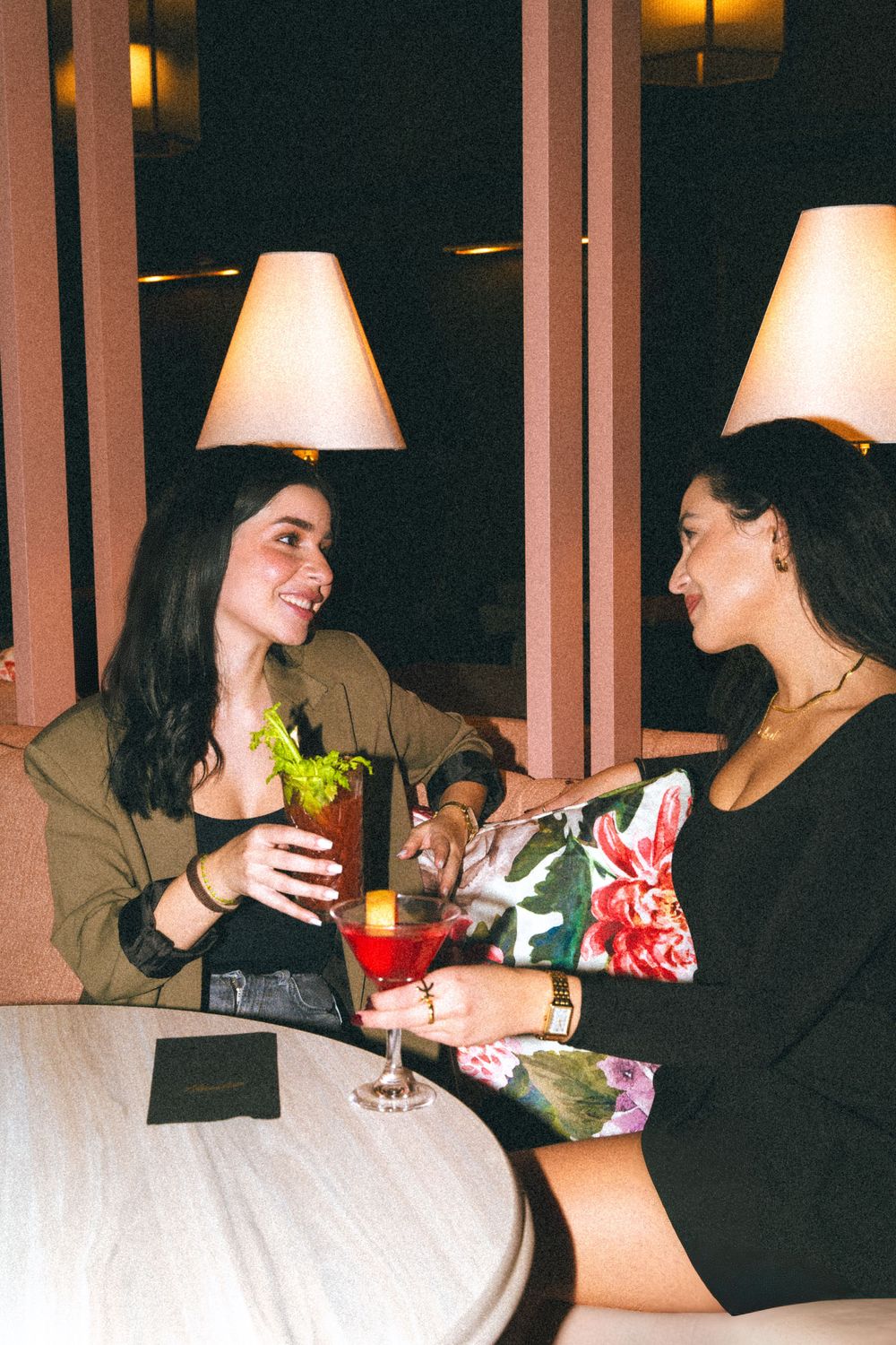 Two friends enjoying cocktails at a dimly lit restaurant with table lamps creating a warm ambiance.