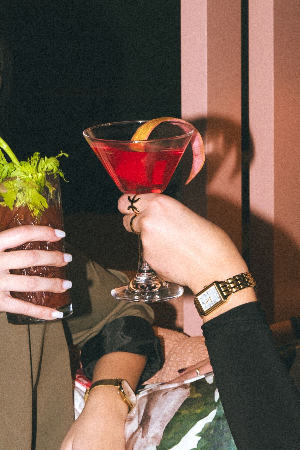Hands holding colorful cocktail glasses with red martini and green garnished drinks at a party against dark background.
