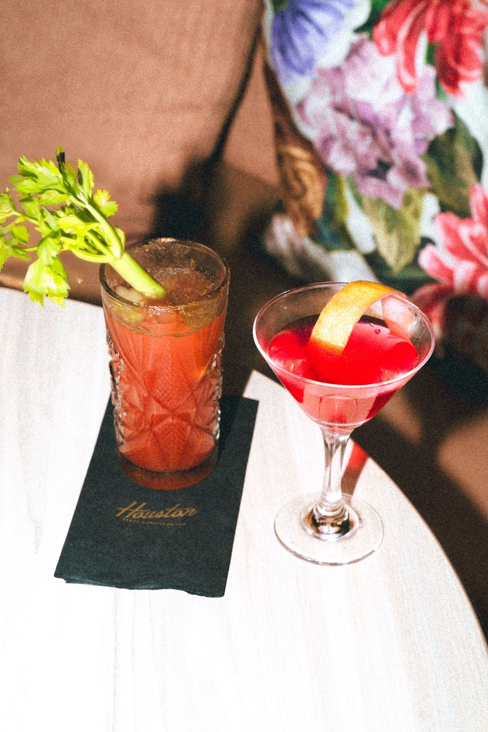 Two colorful cocktails on dark napkins with garnishes, one served in a martini glass and one in a tall glass with celery.