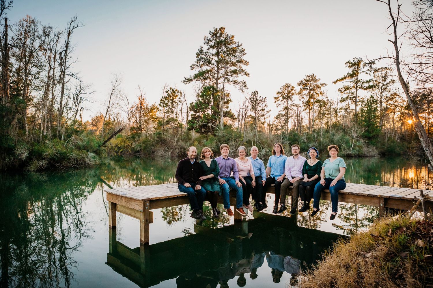 Family group sitting together on wooden dock overlooking tranquil pond at sunset with tall pine trees in background.