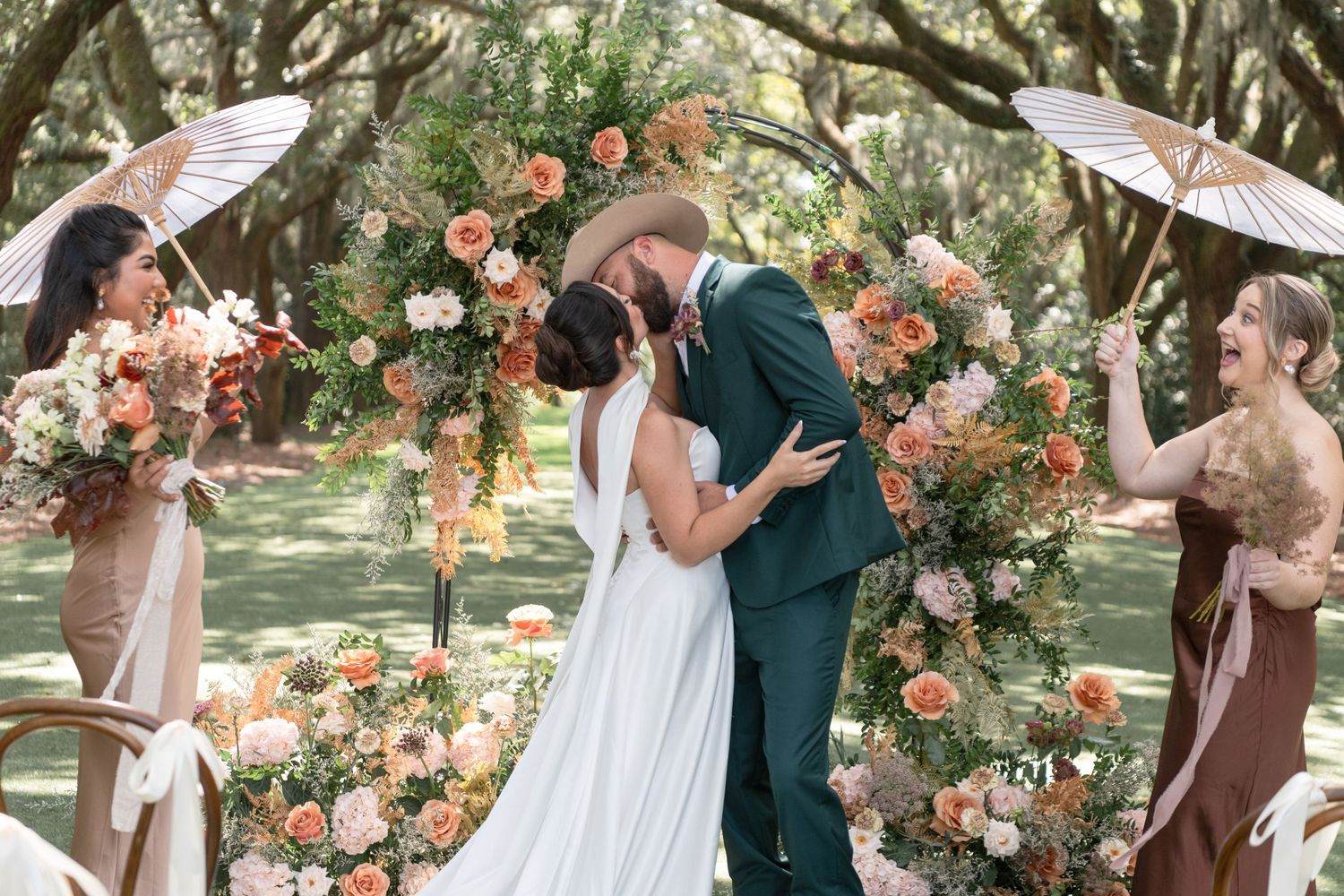 A romantic outdoor wedding ceremony under trees decorated with peach and coral floral arrangements and parasols.