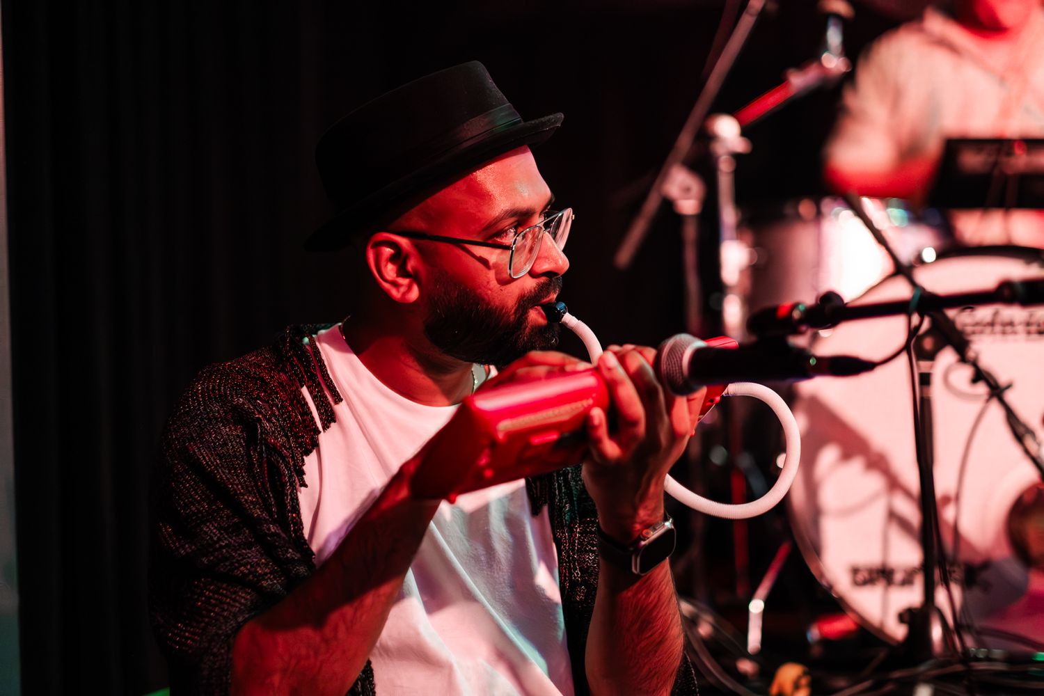 A musician wearing glasses and a black hat performs on stage with a microphone in red-tinted lighting taken by Melbourne event photographer.