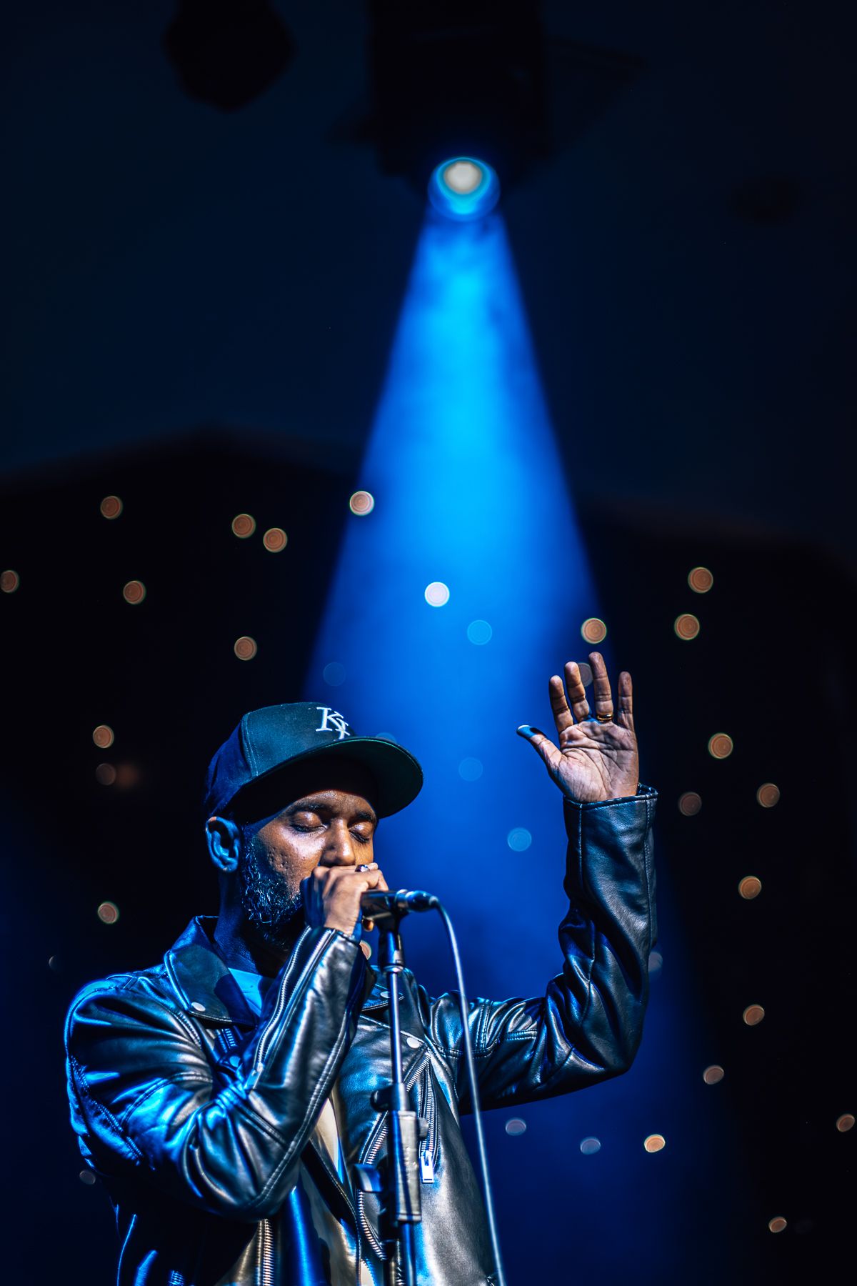 Performer in a leather jacket sings into microphone under dramatic blue spotlight on dark stage taken by Melbourne concert photographer