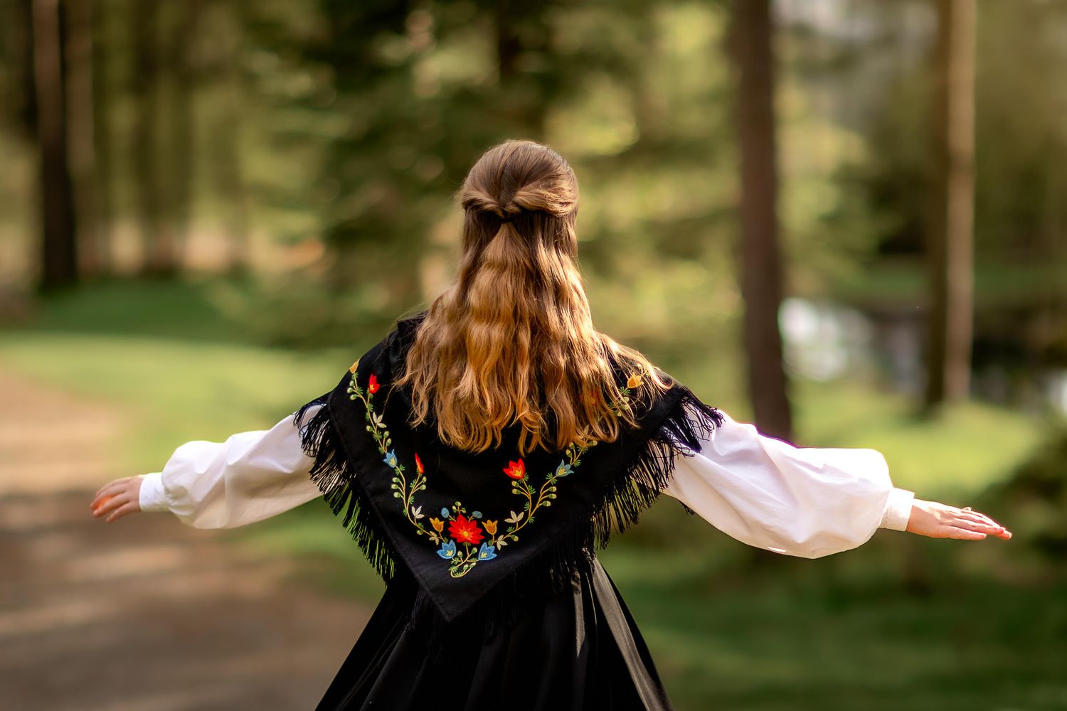 Konfirmant i Frafjordbunad sett bakfra. Hun strekker armene ut og vi ser bølger og sollys i håret hennes.