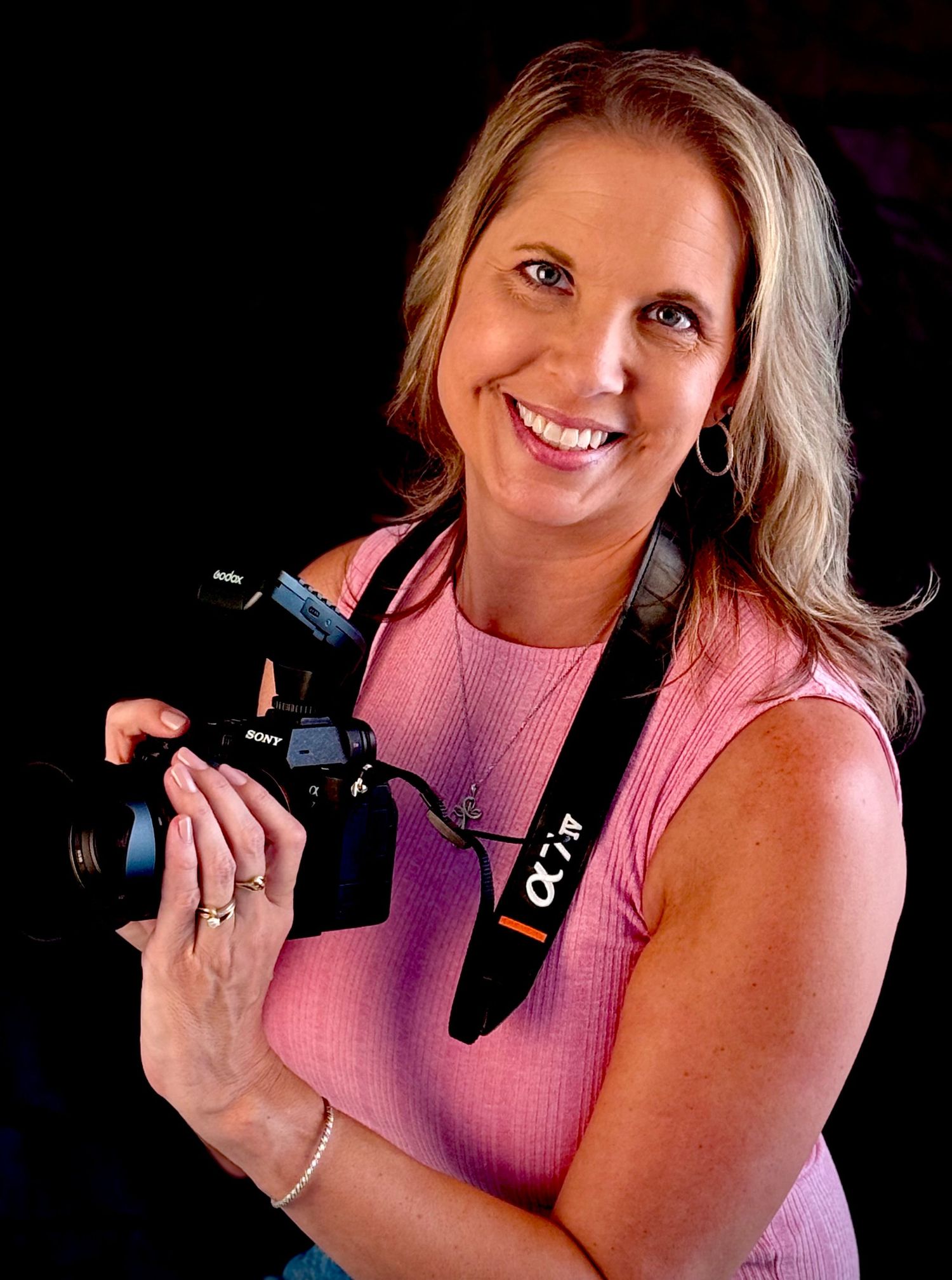 A photographer in a pink top holds a professional camera while smiling against a dark background.