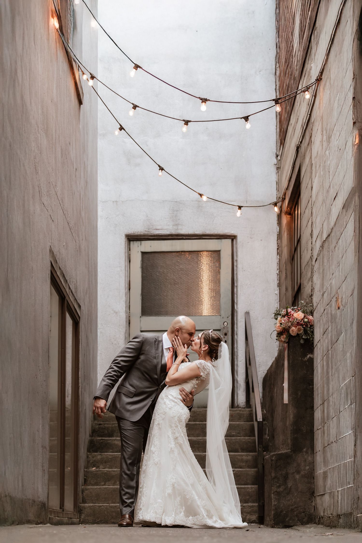 A romantic wedding moment under string lights in an urban alleyway.