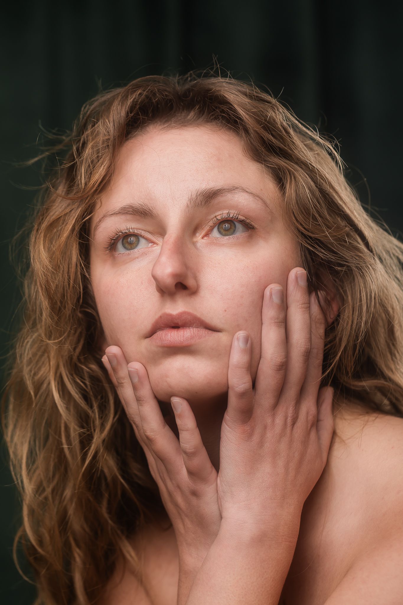 Portrait photograph showing a woman with wavy blonde hair touching her face with both hands against a dark background.