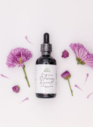 A black dropper bottle surrounded by purple chrysanthemum flowers and petals on a light pink background.