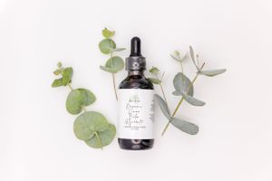 A dark glass dropper bottle with product label lies flat among fresh green eucalyptus leaves on white background.