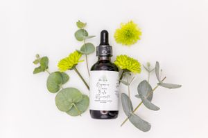 A dark glass dropper bottle with natural beauty product surrounded by yellow flowers and eucalyptus leaves on white background.
