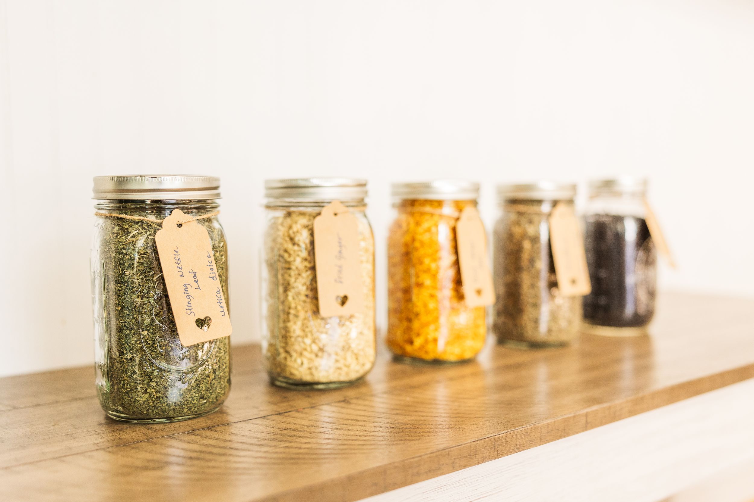 Five glass mason jars filled with colorful spices and herbs arranged in a row on a wooden shelf.