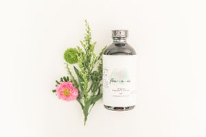 Clear glass bottle with natural skincare product alongside fresh pink flowers and green foliage on white background.