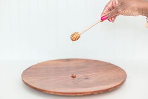 A wooden honey dipper drips honey onto a round wooden plate against a white background.