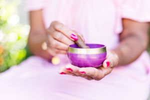 A pair of hands holding a purple meditation singing bowl with manicured pink nails against a soft pink background.