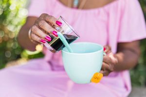 Hot pink manicured hands pour dark liquid into a mint colored teacup with floral accent against blurred green backdrop.