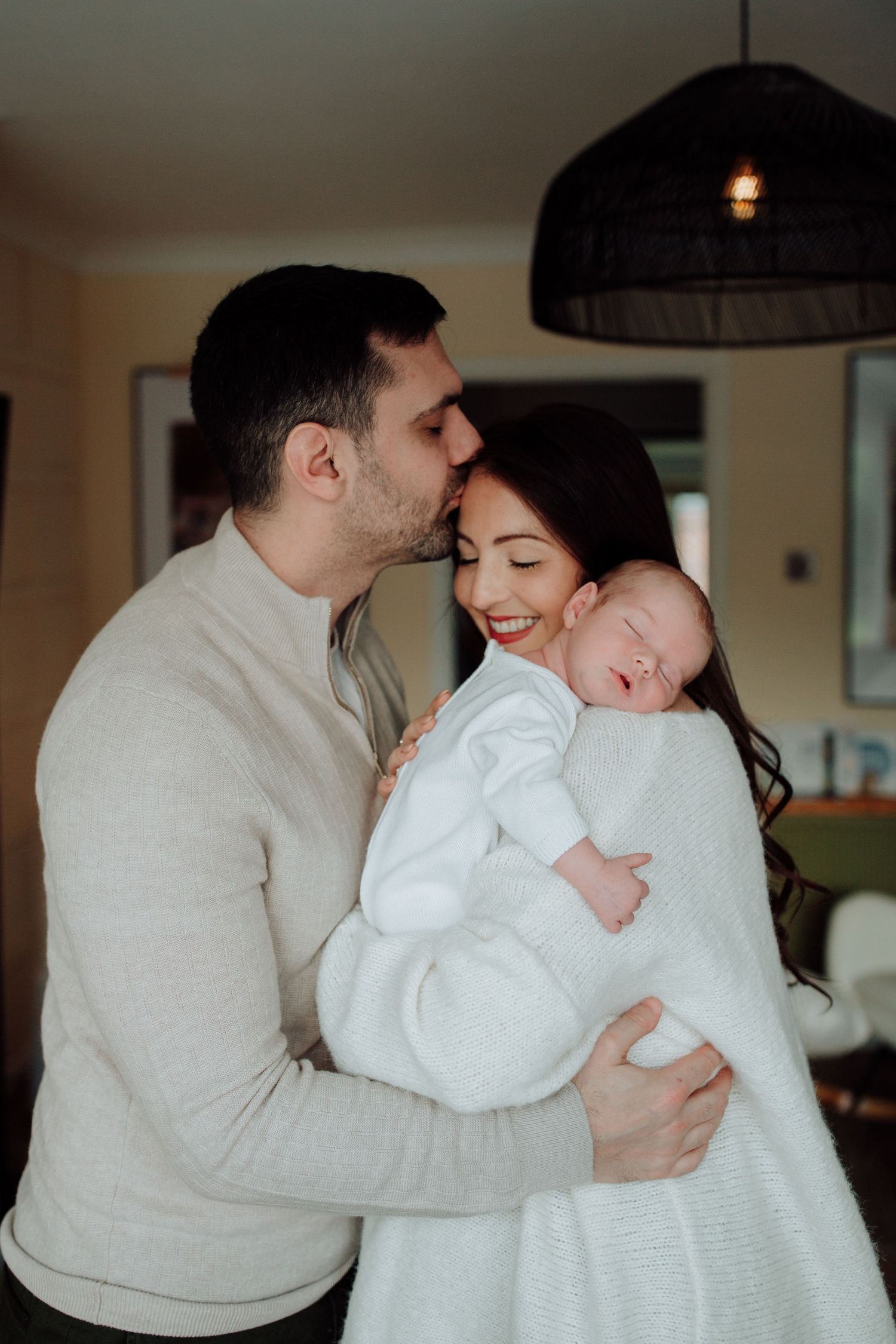 A cozy indoor family moment as parents embrace their sleeping newborn baby wearing white.