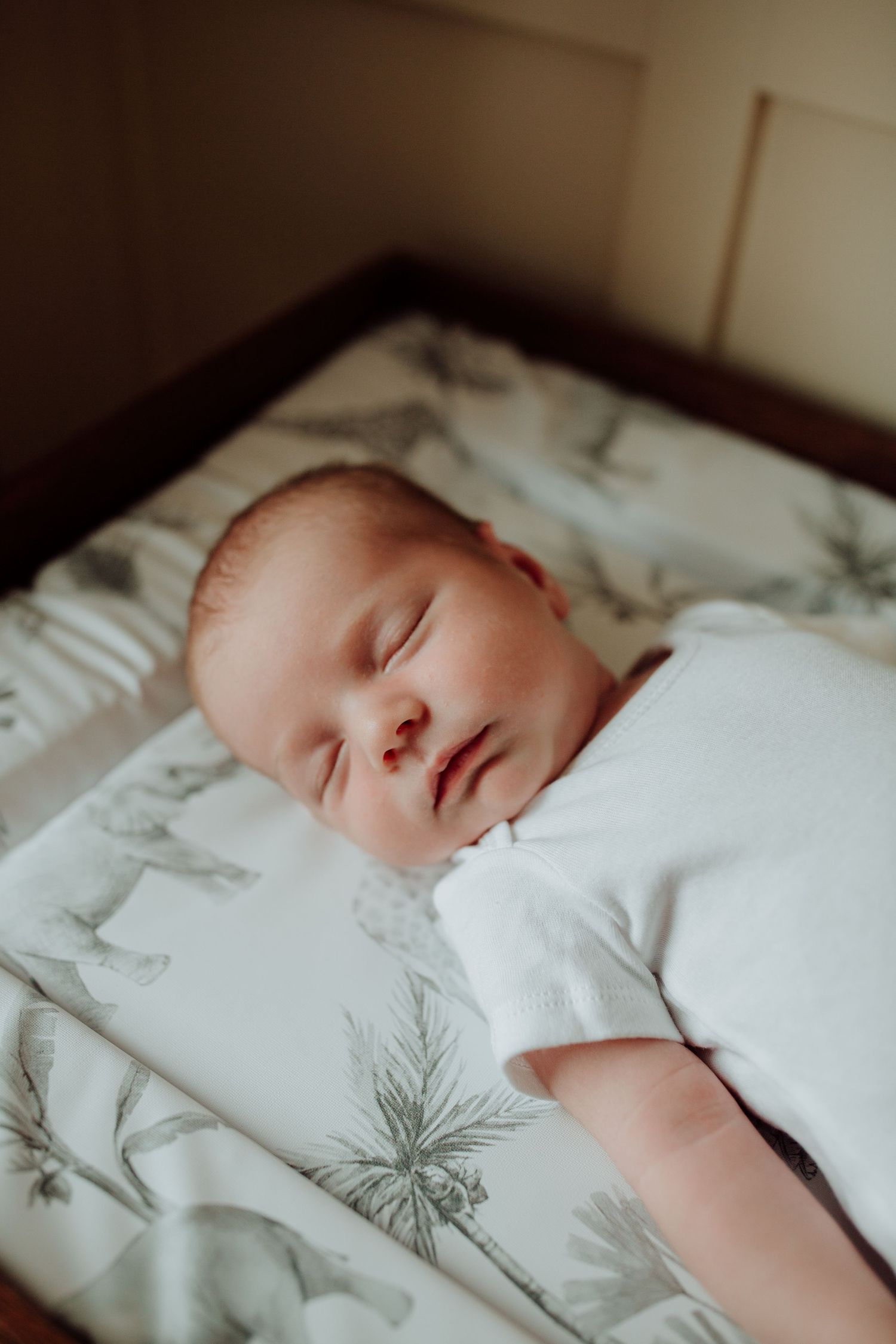 A sleeping infant wearing white lies peacefully on patterned bedding in soft natural light.