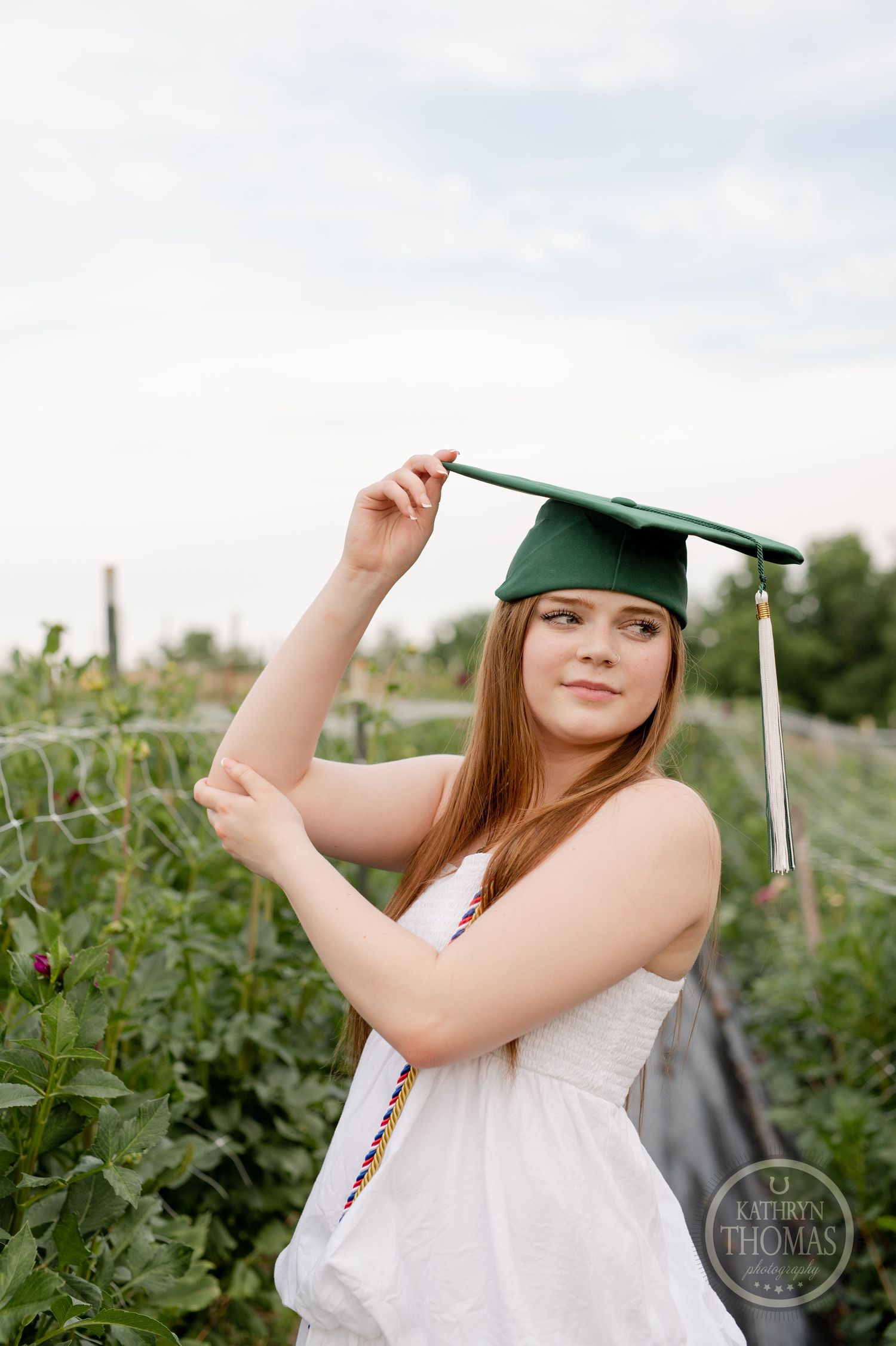 Graduate wearing green cap poses in garden setting with flowers and vines.