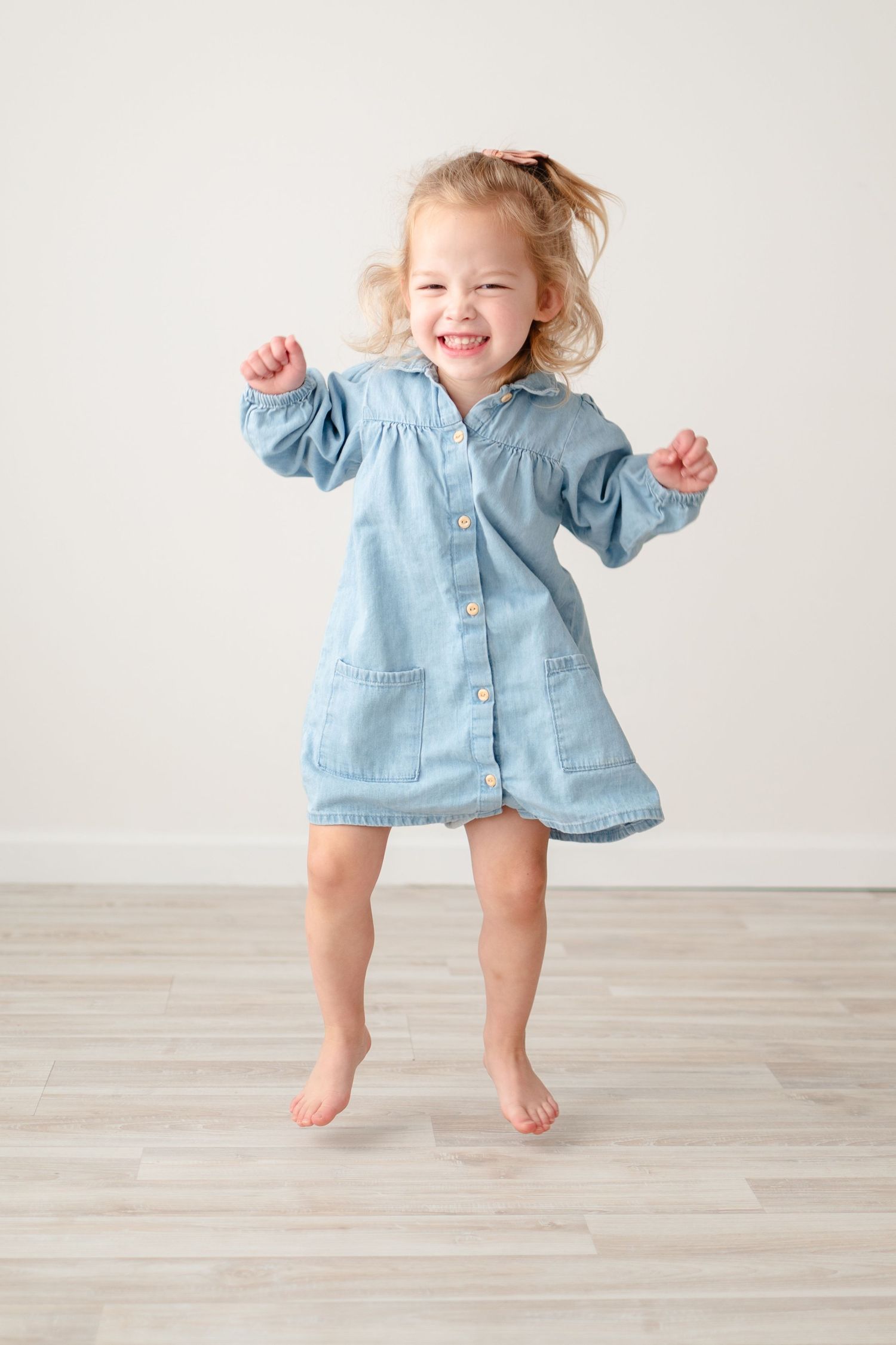 A joyful toddler in a light denim dress jumps and dances barefoot on a light wooden floor against a white wall.
