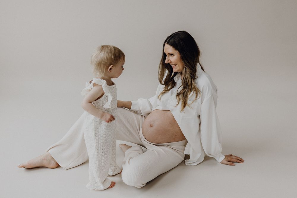 Fotosession im Fotostudio mit einer Schwangeren, diese sitzt auf dem Boden, vor ihr steht ihre Tochter, die auf den bauch blickt.