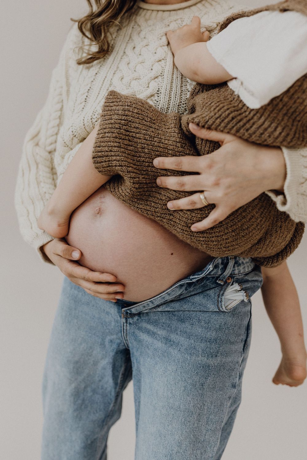 Babybauchshooting im Fotostudio, die Schwangere trägt einen beigefarben Strickpullover, der Bauch ist sichtbar. Auf dem Arm hät sie ein Kind