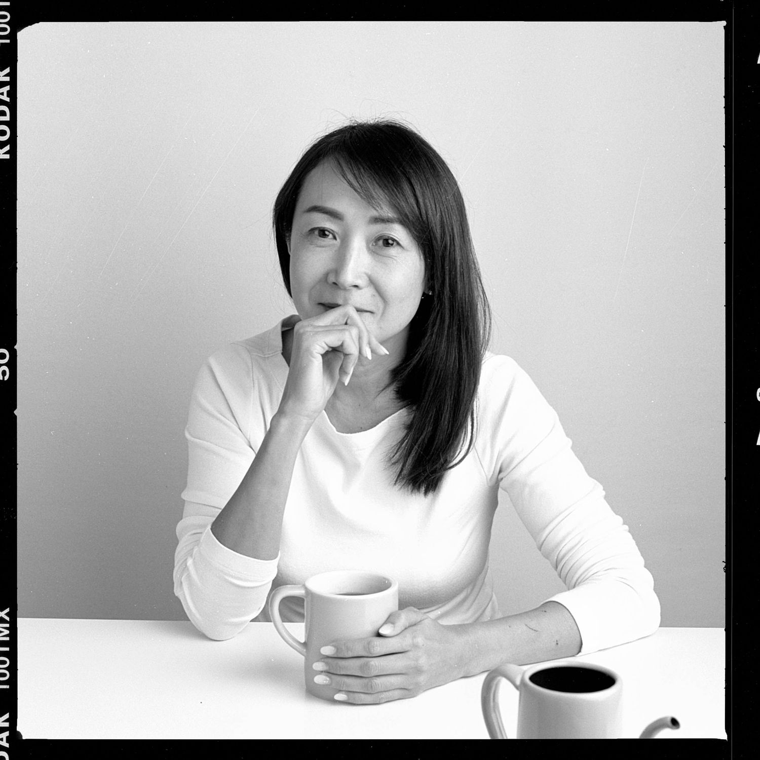 A person in a white long-sleeve shirt sits contemplatively at a table with coffee cups in a black and white photo.