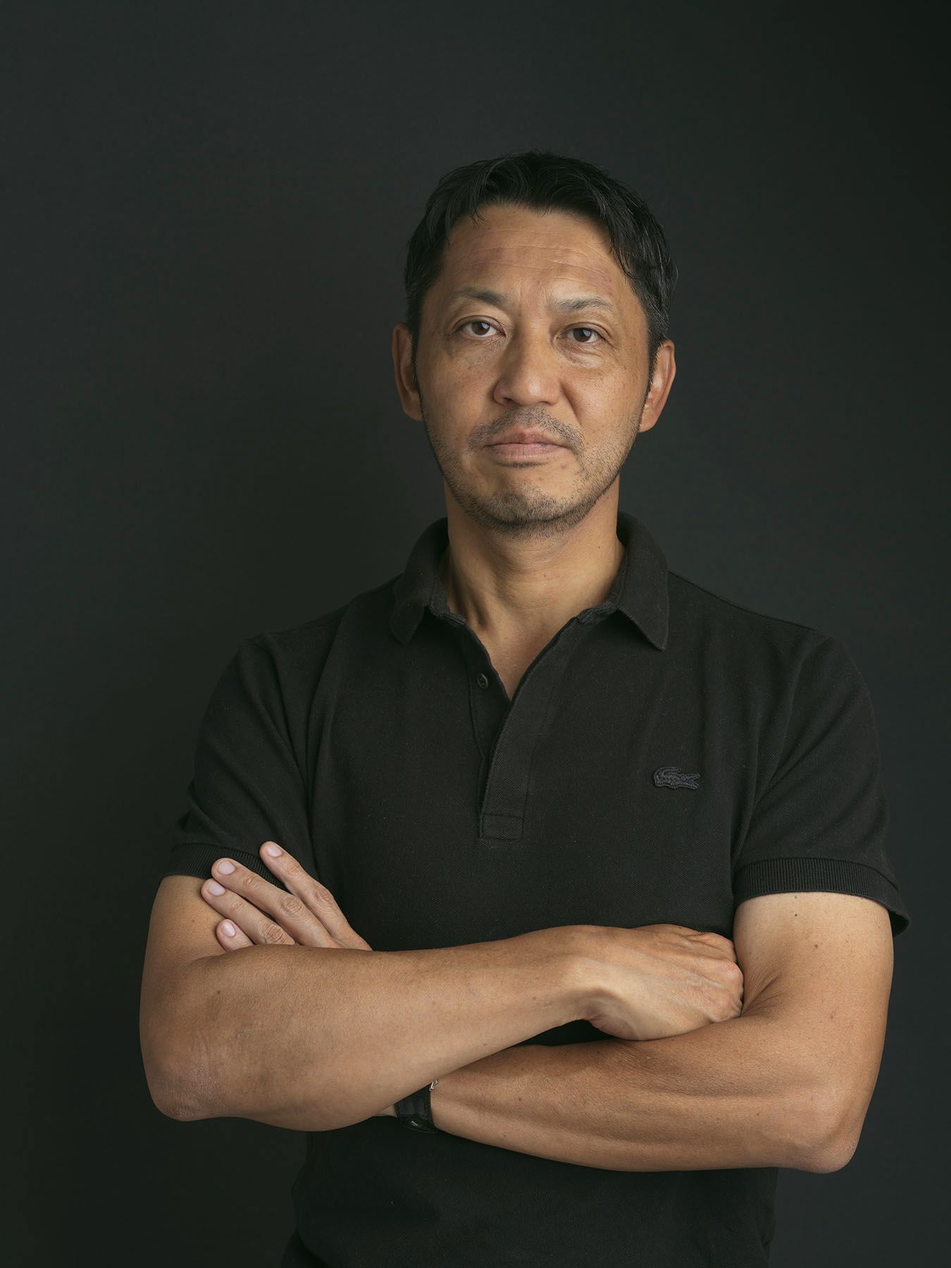 Asian man in black polo shirt stands with arms crossed against dark background in professional portrait.