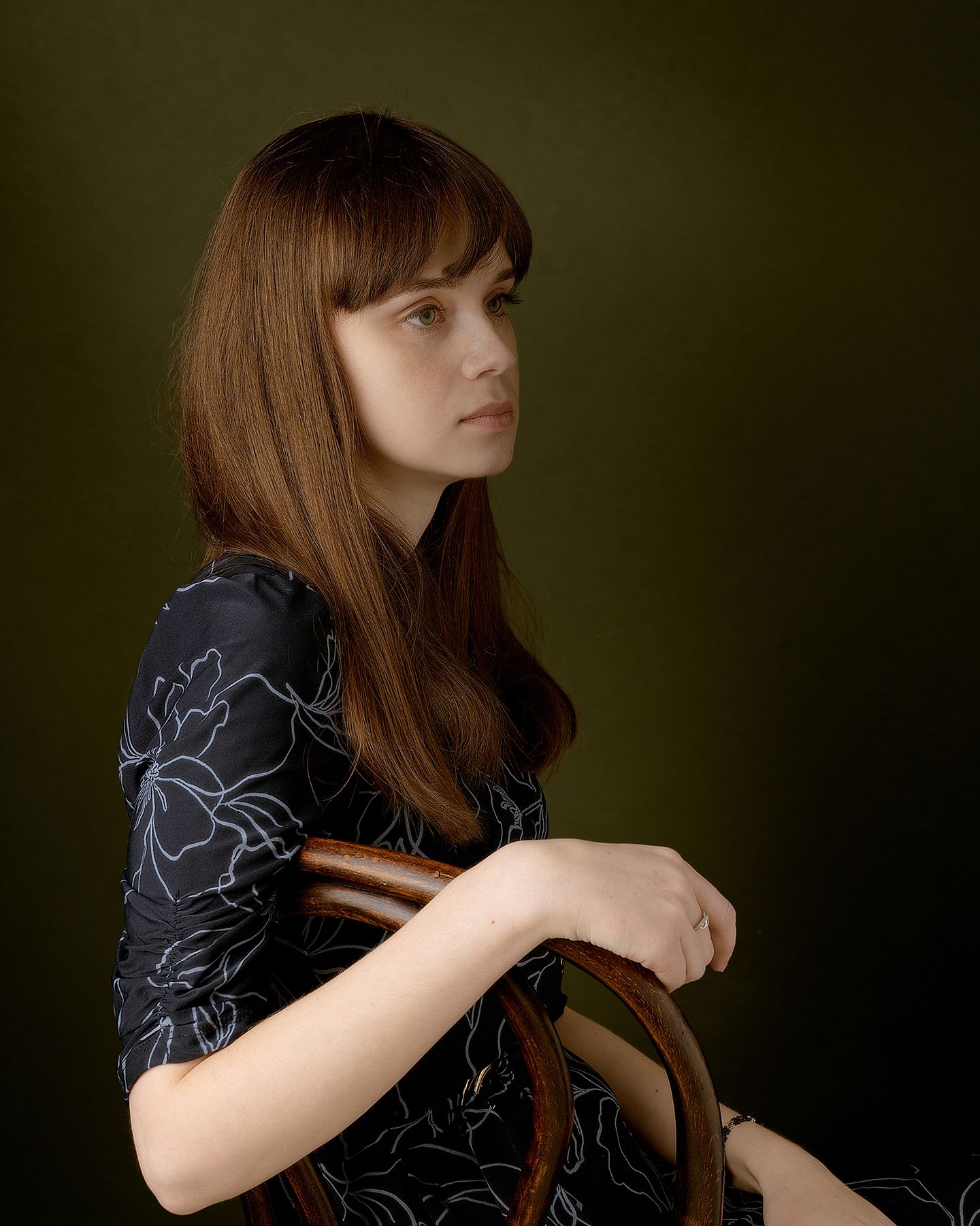 A person in a black patterned dress sits in a dark studio setting with dramatic lighting and long straight hair.