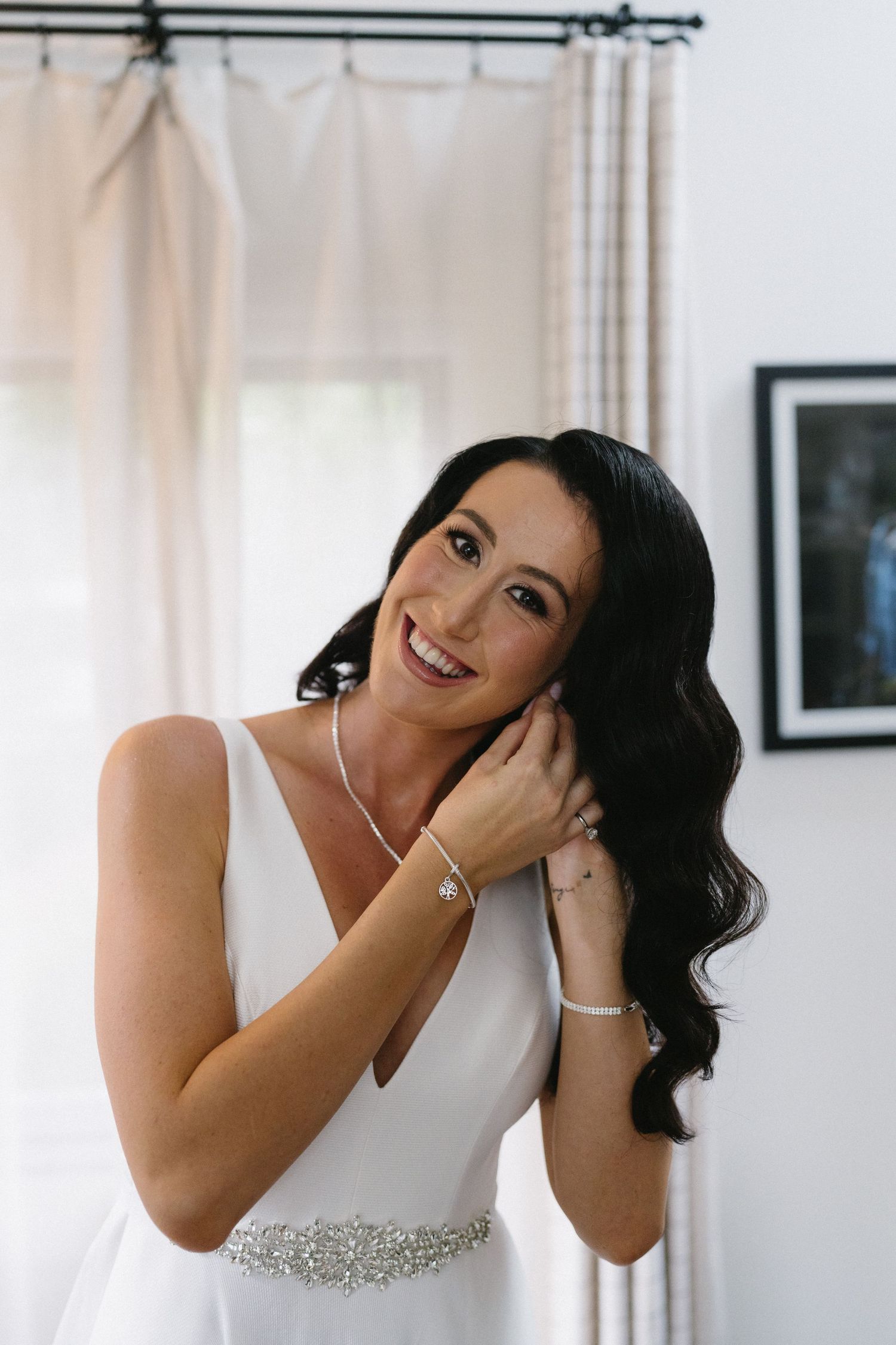 Bride with flowing dark hair touches jewelry while wearing elegant white dress with crystal sash.