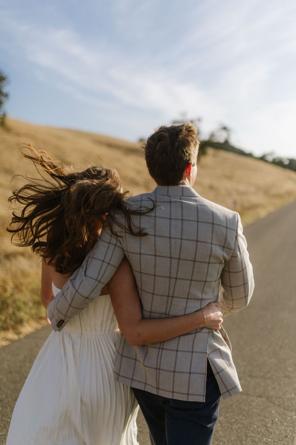 A couple walking away on a winding road with wind blowing through their hair at sunset.