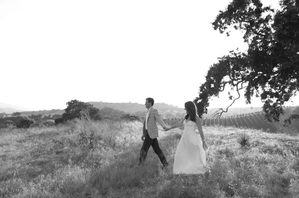 Black and white image of couple walking through scenic hillside meadow in wedding attire.