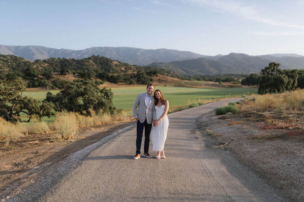 A couple stands together on a winding dirt road surrounded by rolling hills and oak trees in a golden countryside setting.