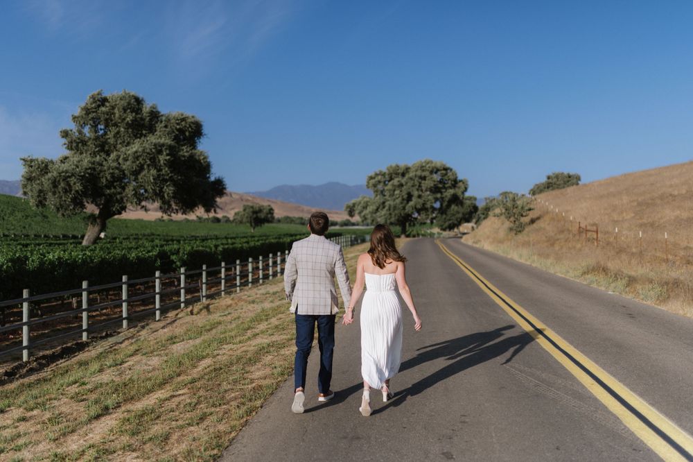 A couple walking hand in hand down a scenic country road with mountains and trees in the background.