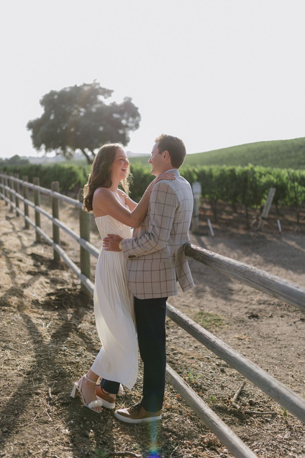 Romantic couple embraces on dirt path along vineyard fence during golden hour sunset.