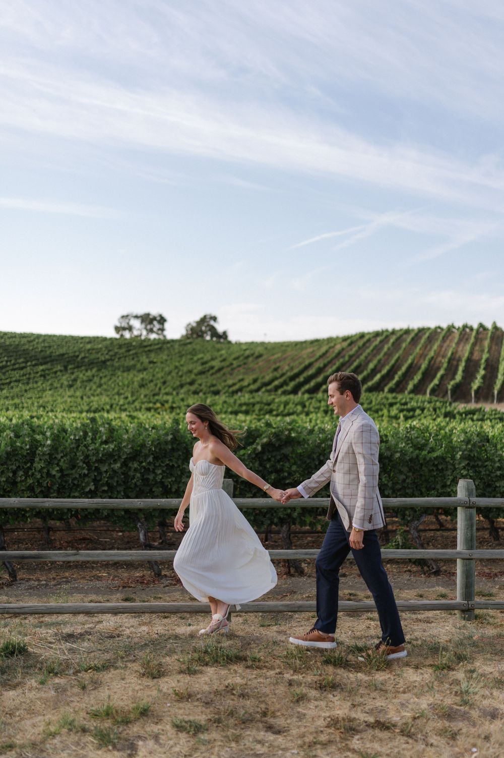 A couple holding hands walks along a fence at a scenic vineyard with rolling hills of grapevines under a cloudy sky.