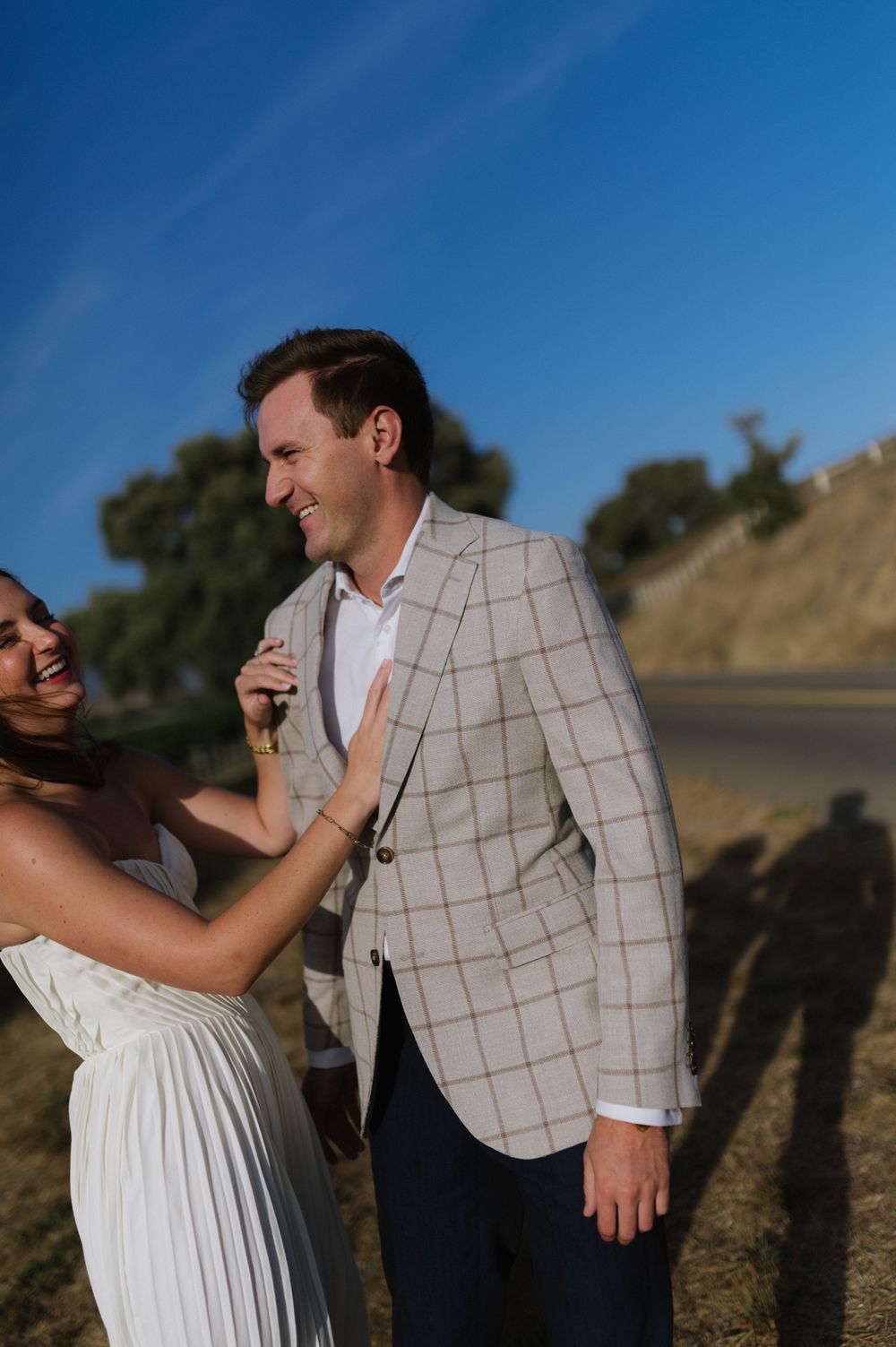 A romantic moment captured of a couple in white and plaid laughing together against a sunny hillside backdrop.
