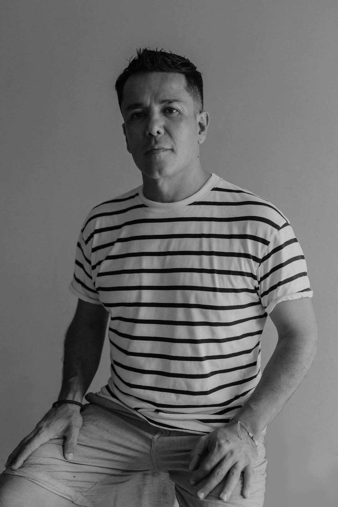 Person wearing a black and white striped t-shirt sitting on a stool against a plain backdrop.
