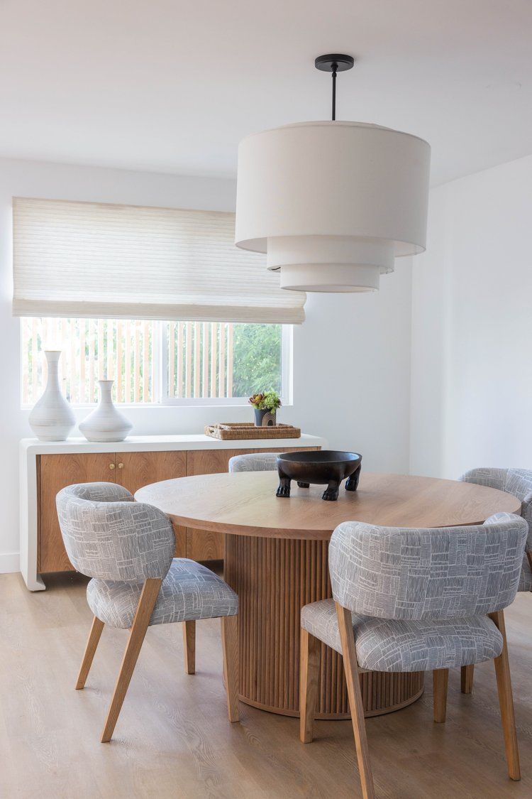 Modern dining area with round wooden table, upholstered gray chairs, white drum pendant light and bamboo window shades.