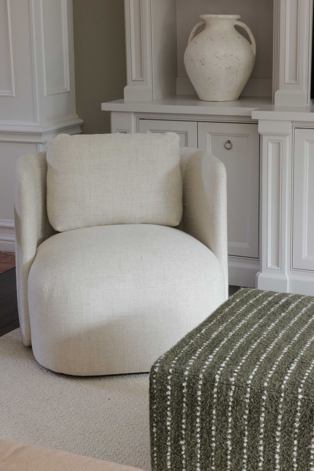Modern cream swivel accent chair with curved design situated next to a green striped ottoman beneath a built-in shelf display.