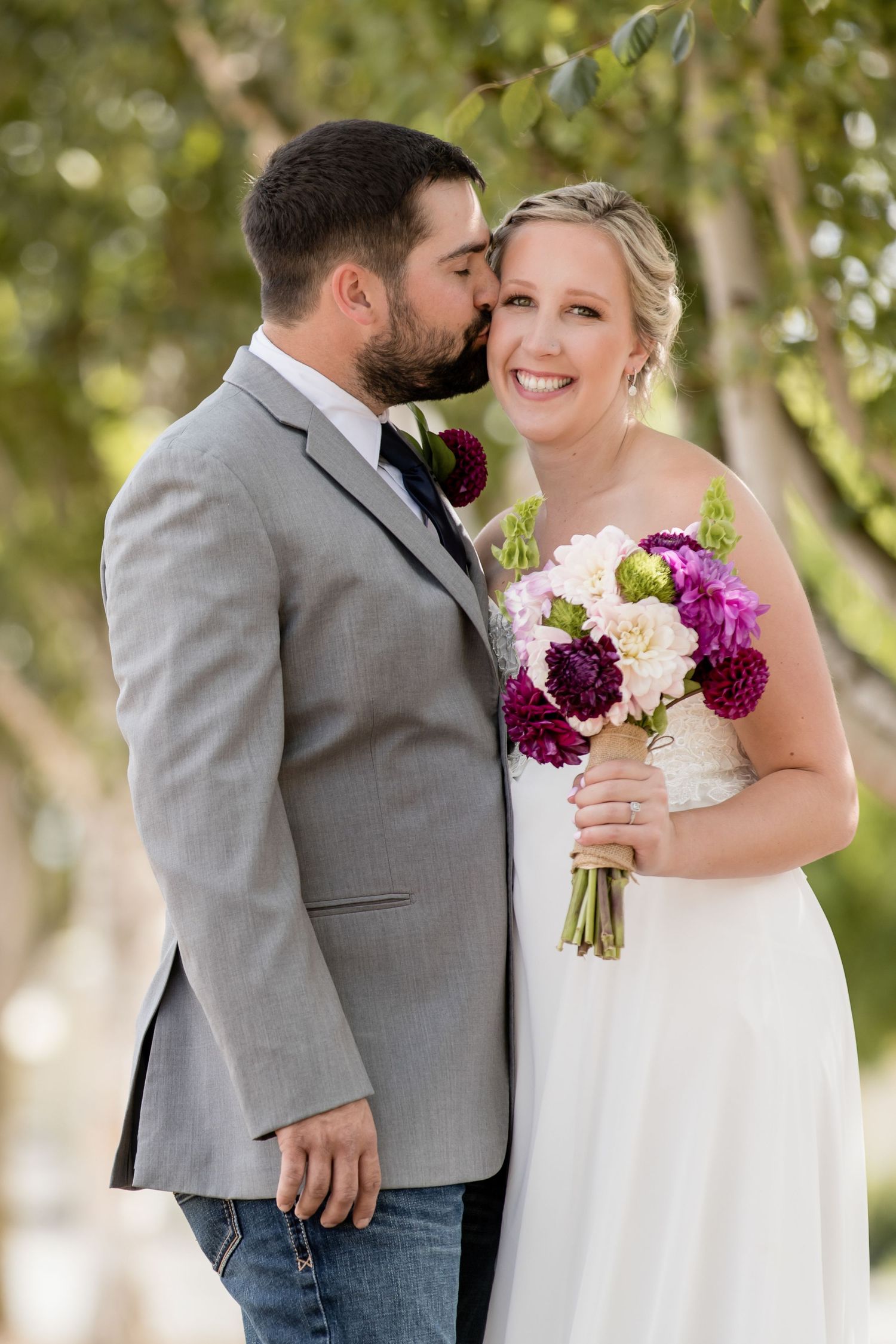 Newlyweds share tender moment outdoors with purple and white floral bouquet.