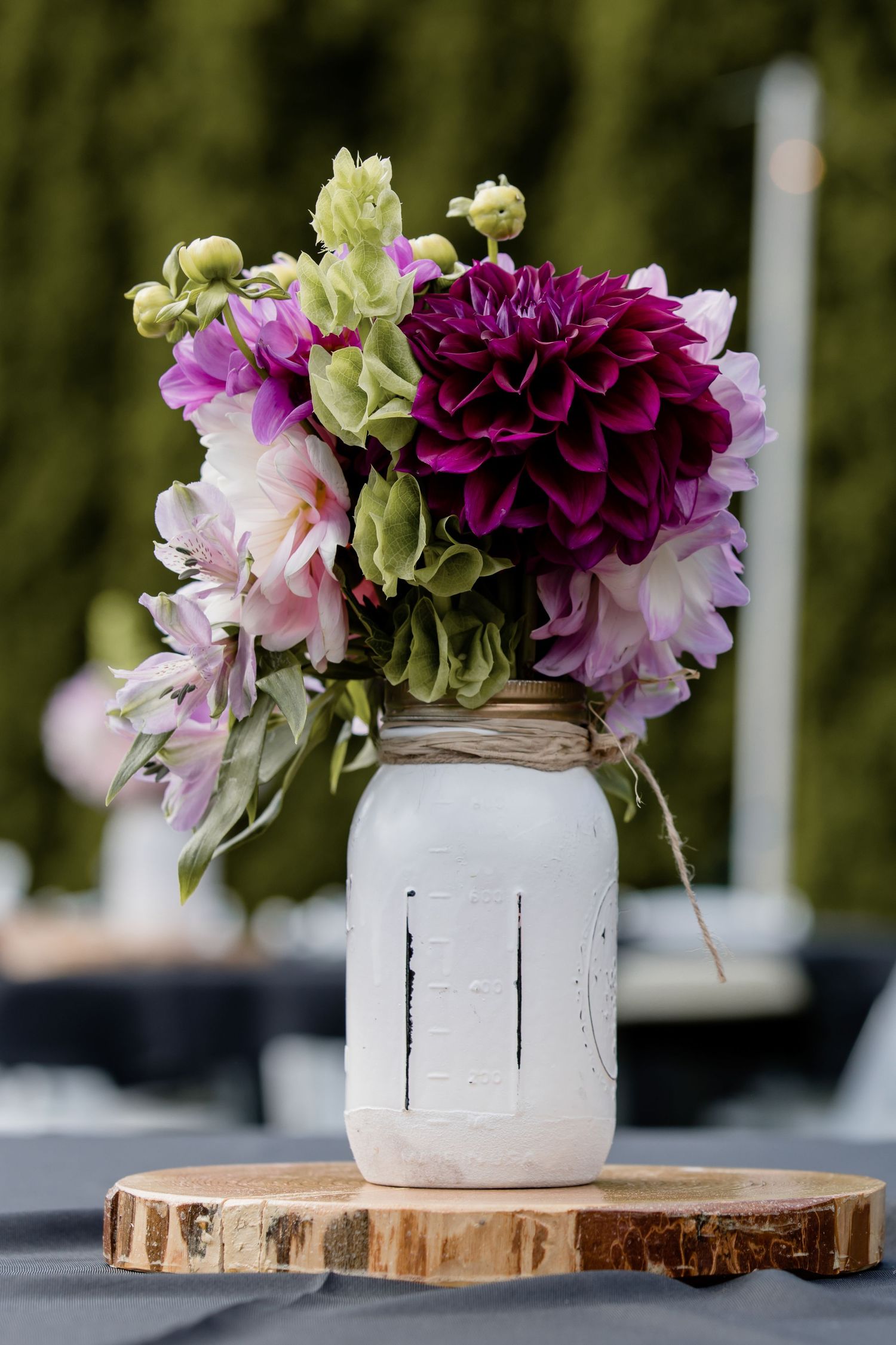 Purple dahlia and pink flower arrangement in white painted mason jar on wooden slice centerpiece.