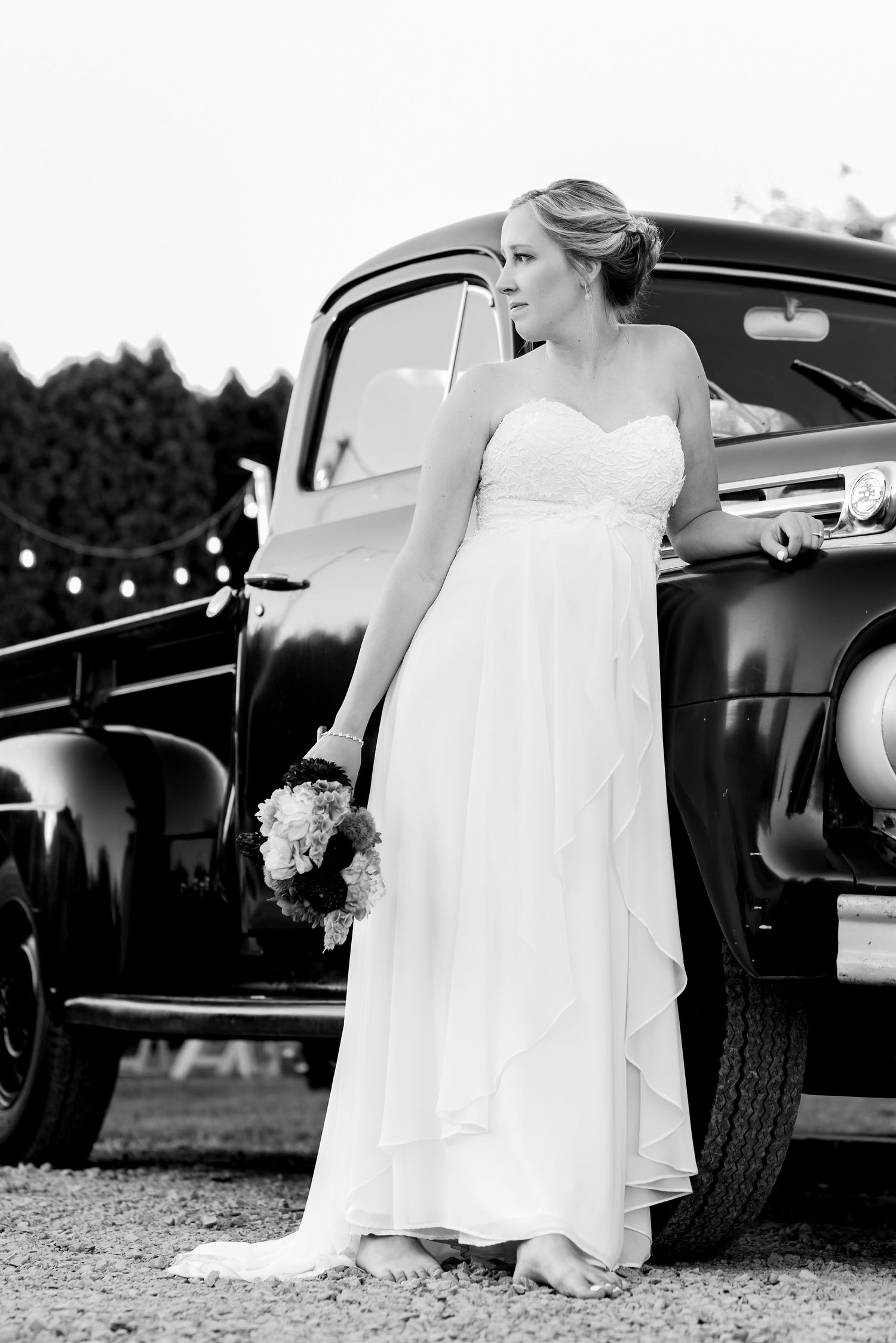 Classic black and white portrait of bride in flowing dress posed against vintage truck.