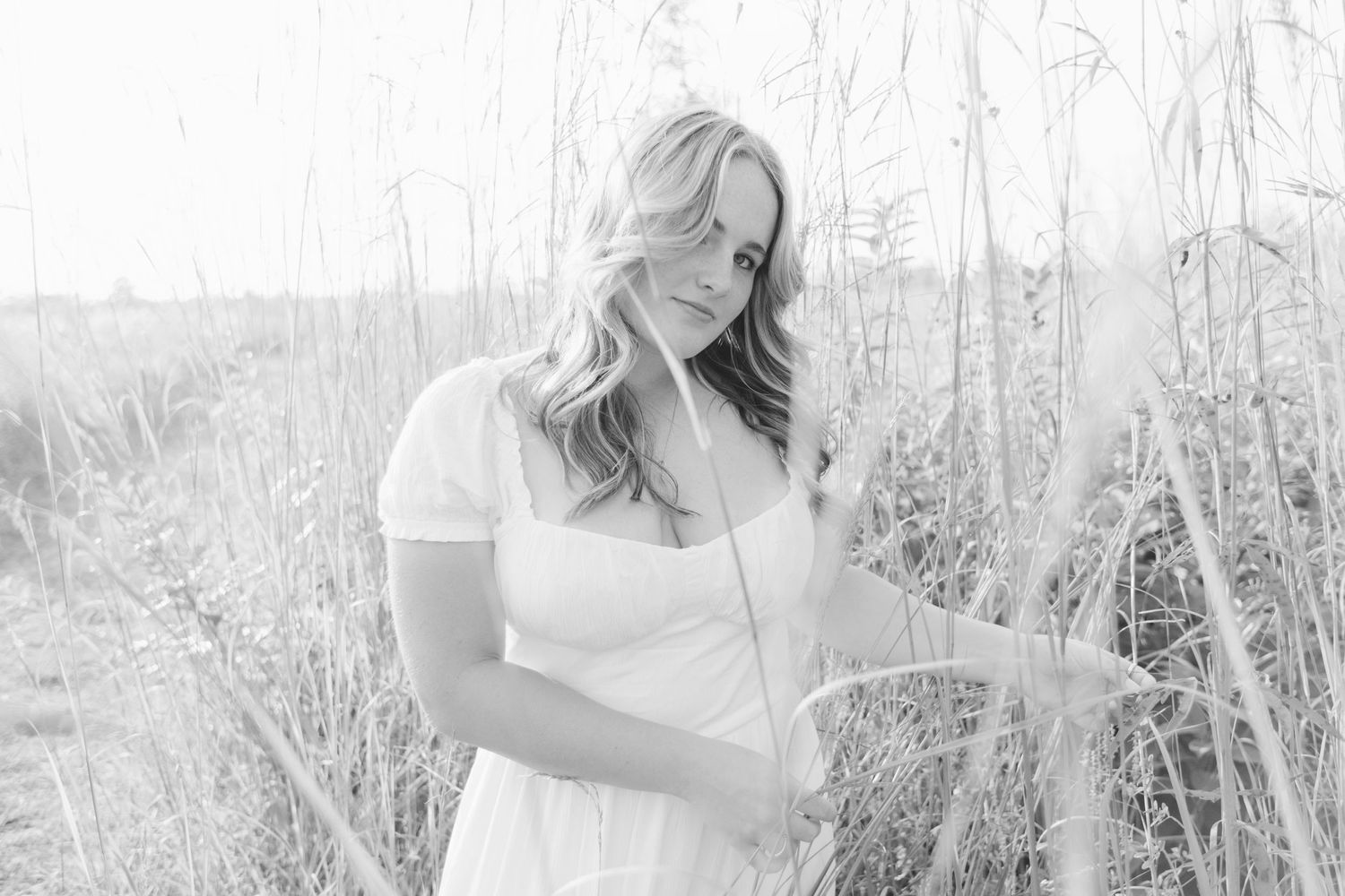 Black and white portrait of a person in a white dress standing thoughtfully among tall grasses in a field.