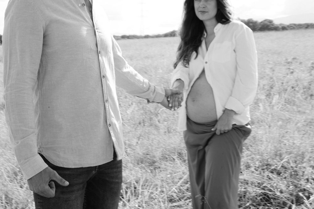 Black and white photo of couple holding hands in field during maternity shoot.