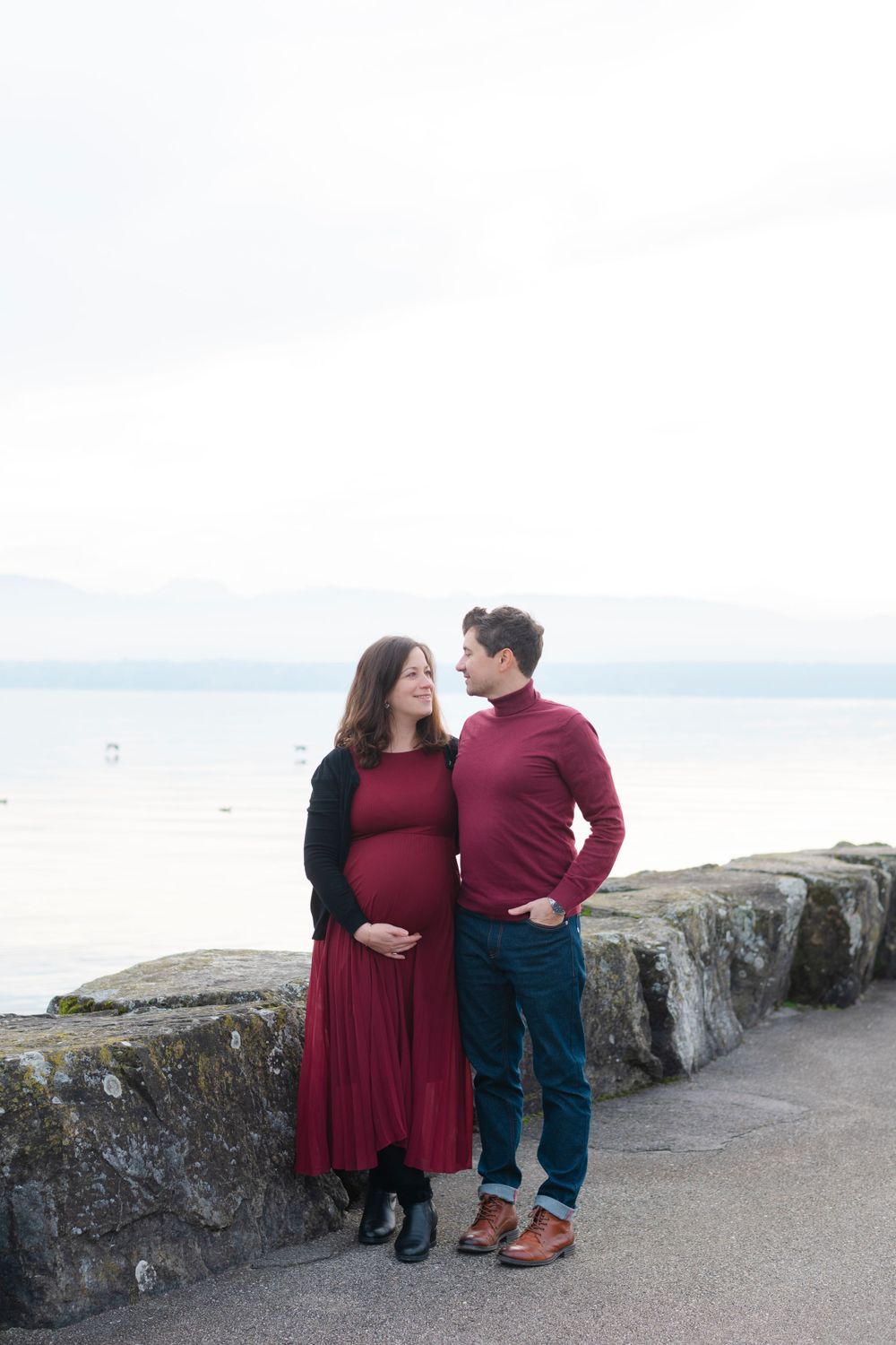 Couple standing by stone wall overlooking water in matching burgundy outfits.