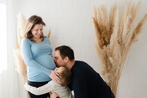 An expectant couple sharing a tender moment in front of dried pampas grass against a white wall.