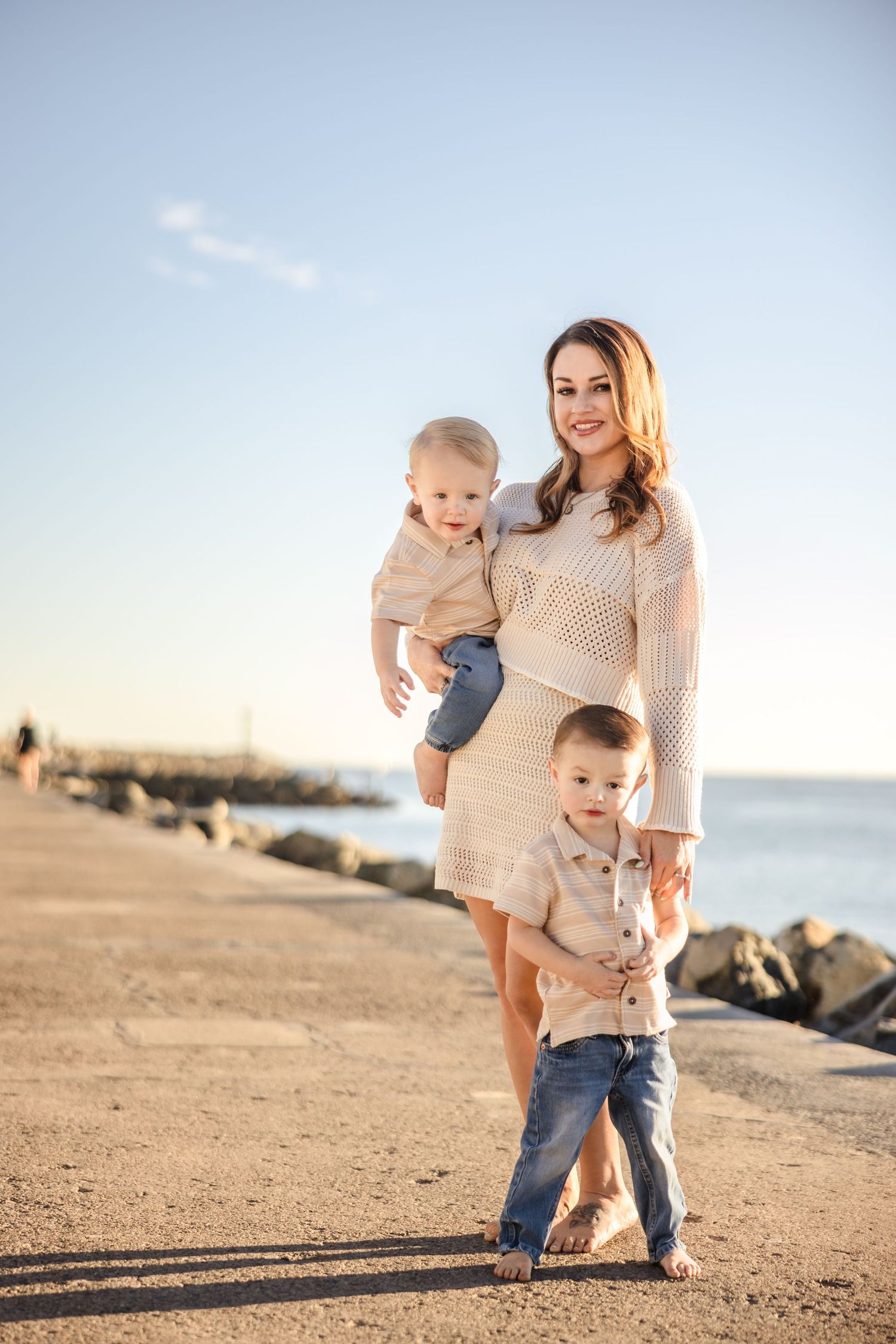 A family poses together on a sandy beach near rocky shores at sunset wearing coordinated beige sweaters and jeans.