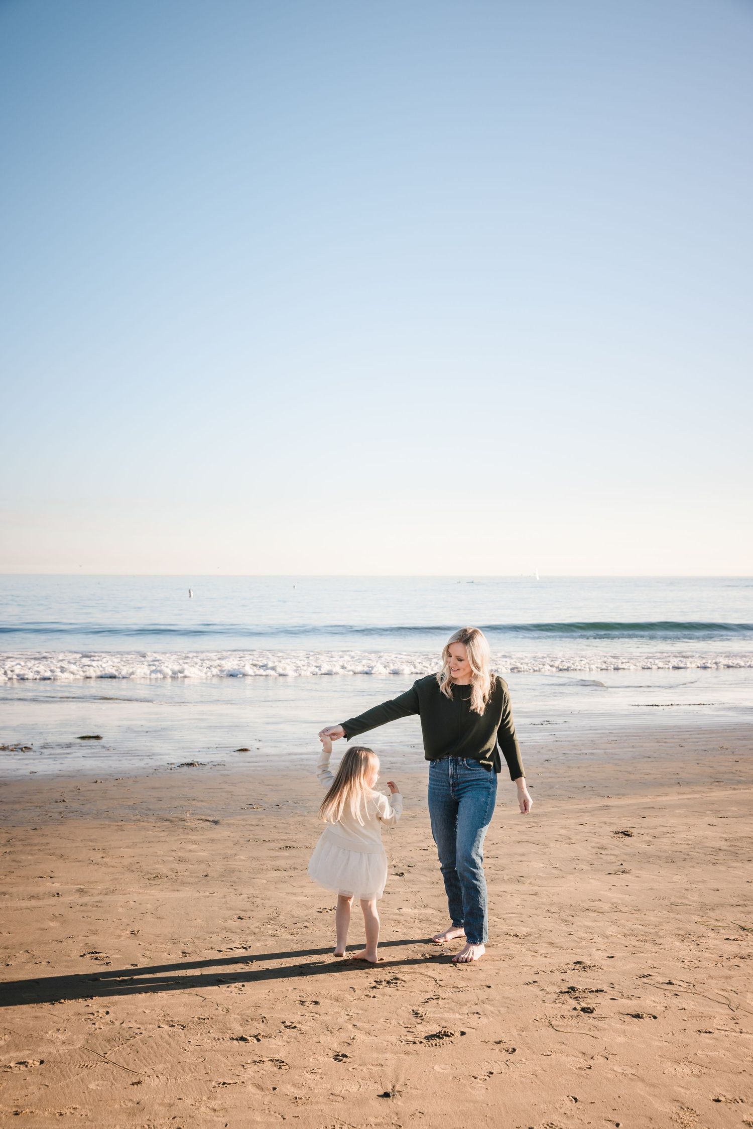 Two people walking together and holding hands on a sunny beach with calm ocean waves in the background.